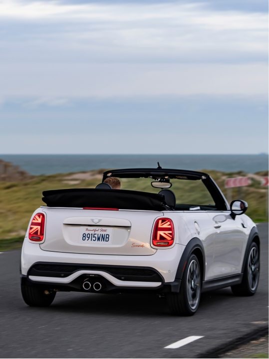 Three-quarters back view of a MINI Cooper S Convertible Seaside Edition in Nanuq White parked on dark pavement with its top down along with a grassy hill and cloudy skies in the background.