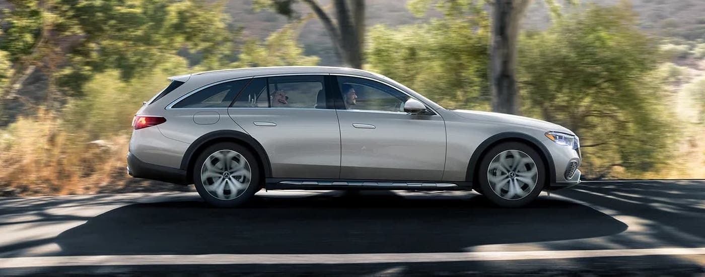 A silver 2024 Mercedes-Benz E-Class Wagon is driving on a tree-lined road.