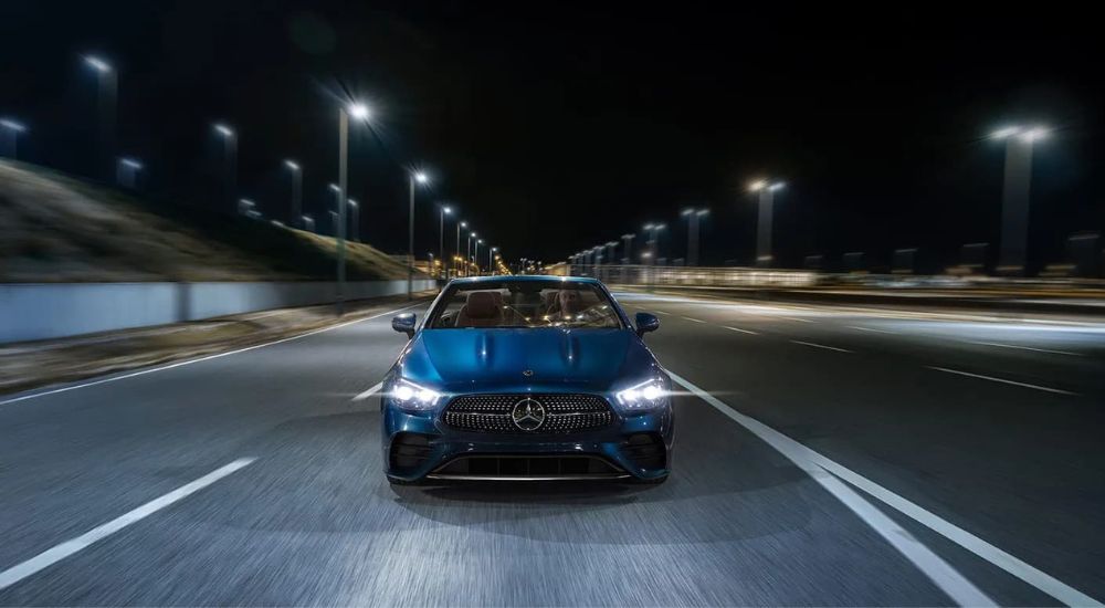 A popular used Mercedes-Benz for sale in Medford, a blue 2022 Mercedes-Benz E-Class, is shown driving on a city street at night.