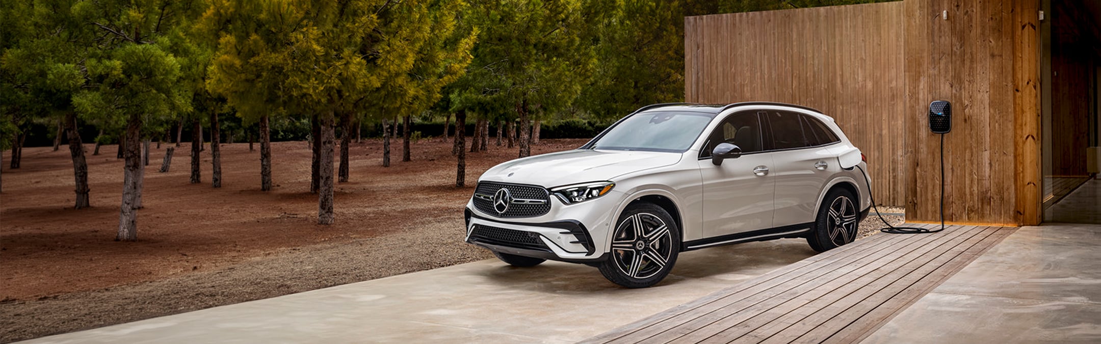 A white Mercedes-Benz GLC plugged into a wall-mounted home charger beside a wooden building