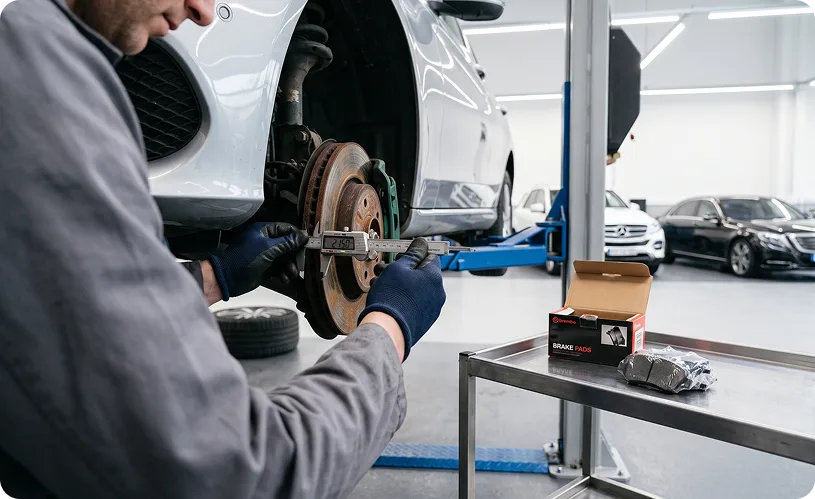 Technician measuring the thickness of a worn brake rotor with digital calipers on a silver Mercedes-Benz, with a boxed set of replacement brake pads on the bench beside him