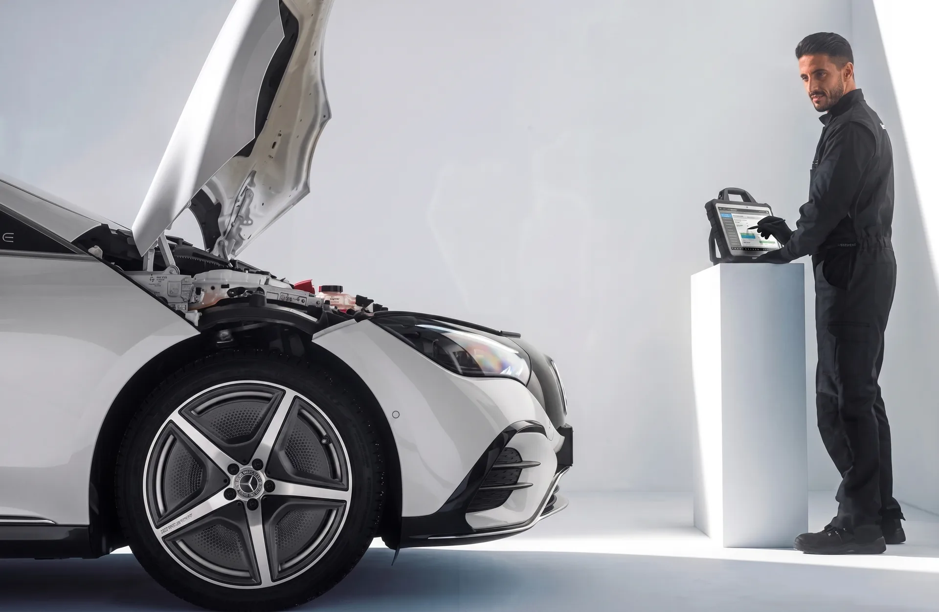 A technician in dark overalls working at a diagnostic tablet beside a white Mercedes-Benz with the hood raised