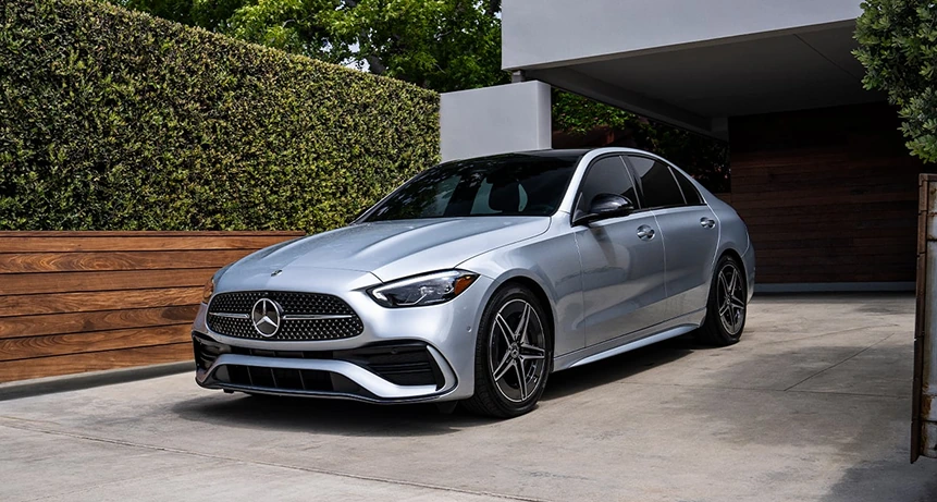 Silver C-Class Sedan with AMG Line styling parked in a driveway beside a hedge and wooden fence