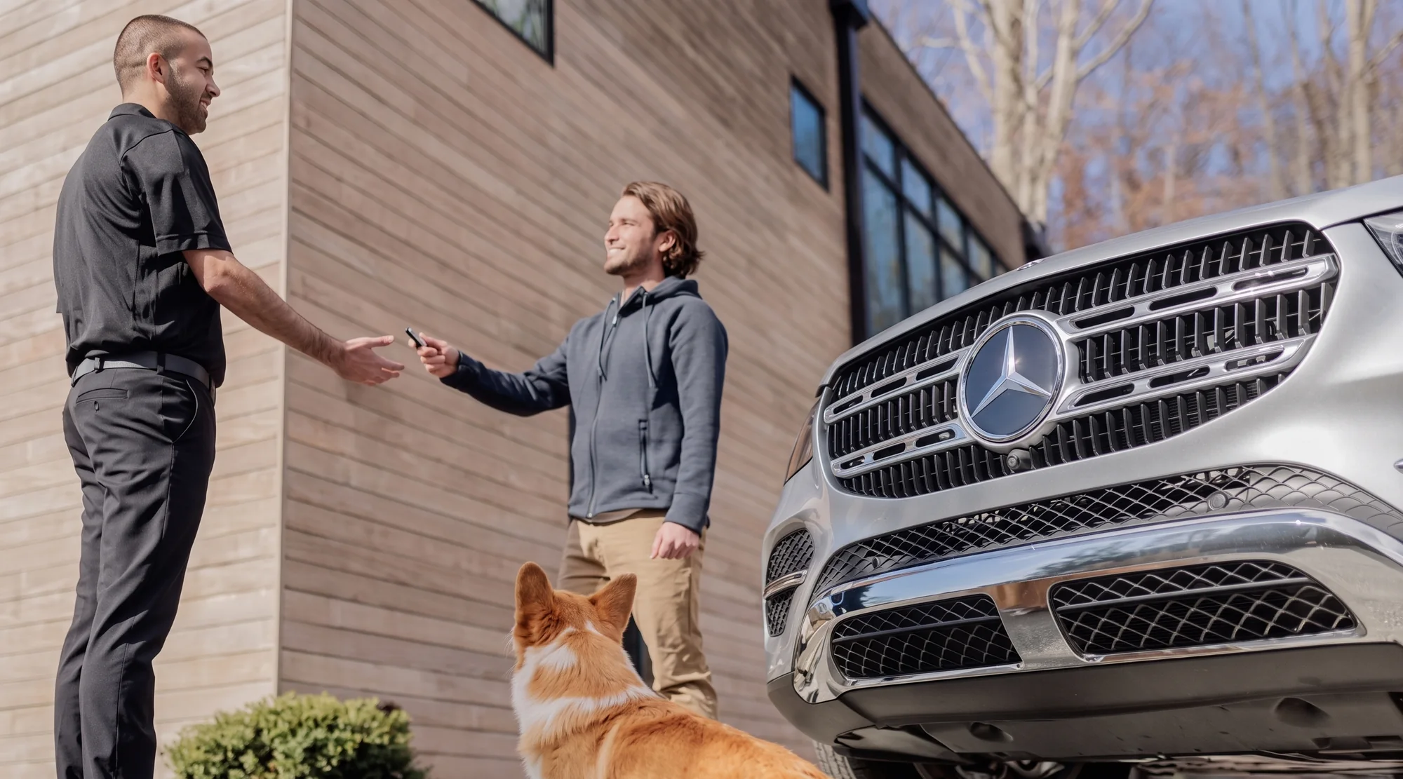 A Mercedes-Benz team member in a black polo handing a key fob to a customer beside a silver SUV outside a house, a corgi sitting between them