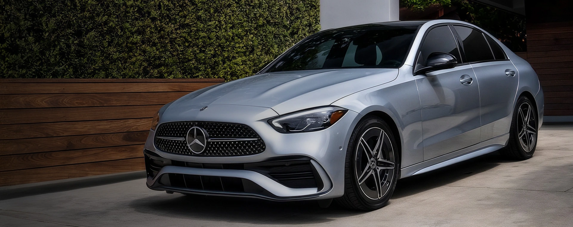 Silver 2026 Mercedes-Benz C-Class Sedan parked on a paved drive in front of a wooden fence and a tall green hedge