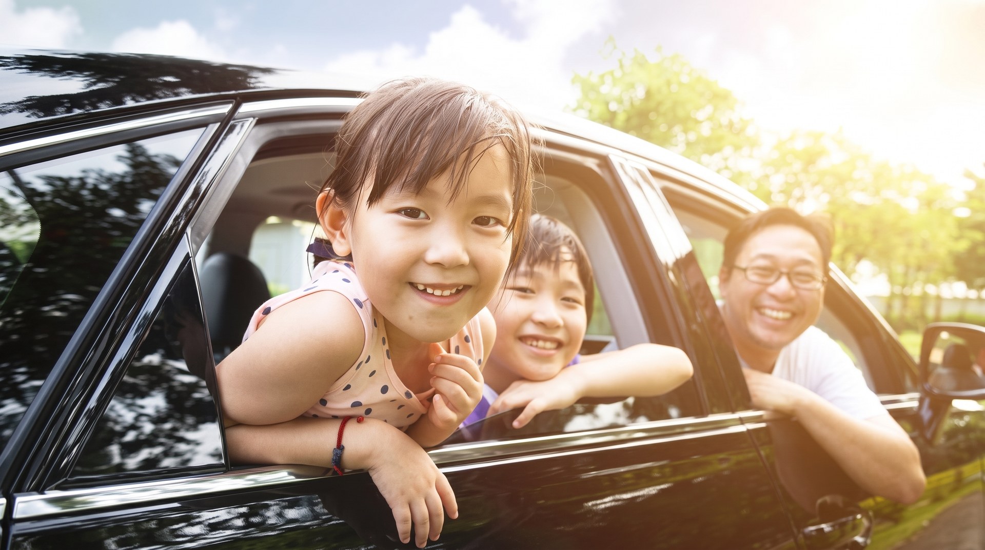 Two children leaning out of a car window on a sunny day, with an adult beside them