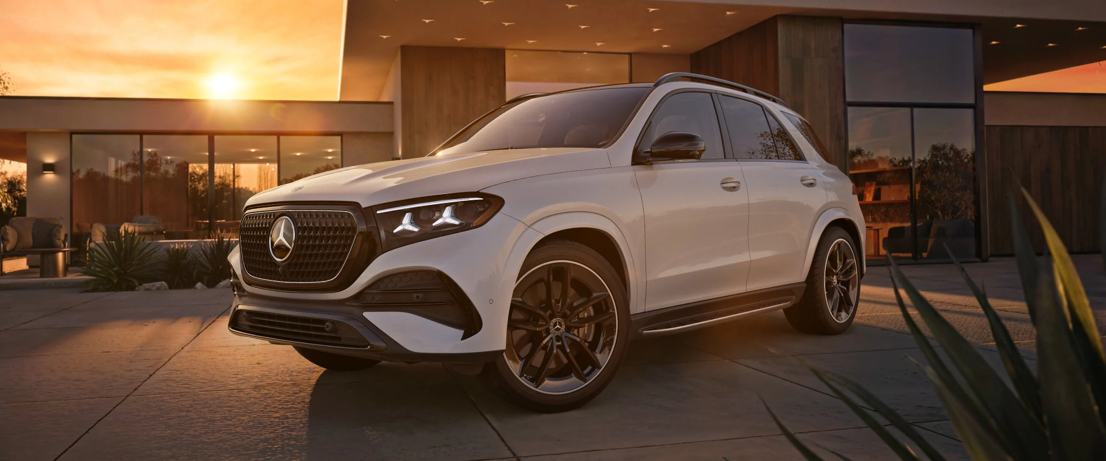 A white Mercedes-Benz GLE SUV parked on the drive of a modern house at sunset, front three-quarter view