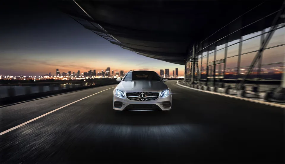 A silver Mercedes-Benz E-Class coupe driving toward the camera on a bridge at dusk, the city skyline lit behind it