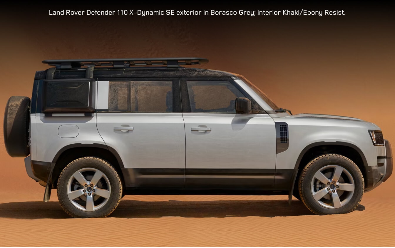 Side profile view of a 2026 Land Rover Defender 110 X-Dynamic SE in Borasco Grey equipped with the Explorer Pack parked on desert sand dunes.
