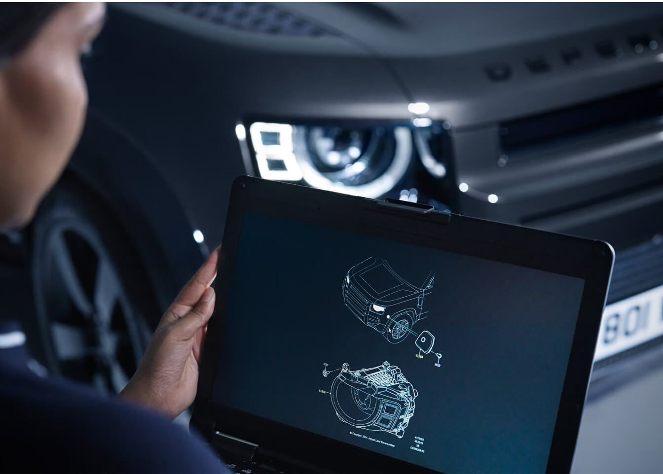 Over-the-shoulder view of a Land Rover certified technician reviewing diagnostic schematics on a laptop in front of a 2026 Land Rover Defender 110 .