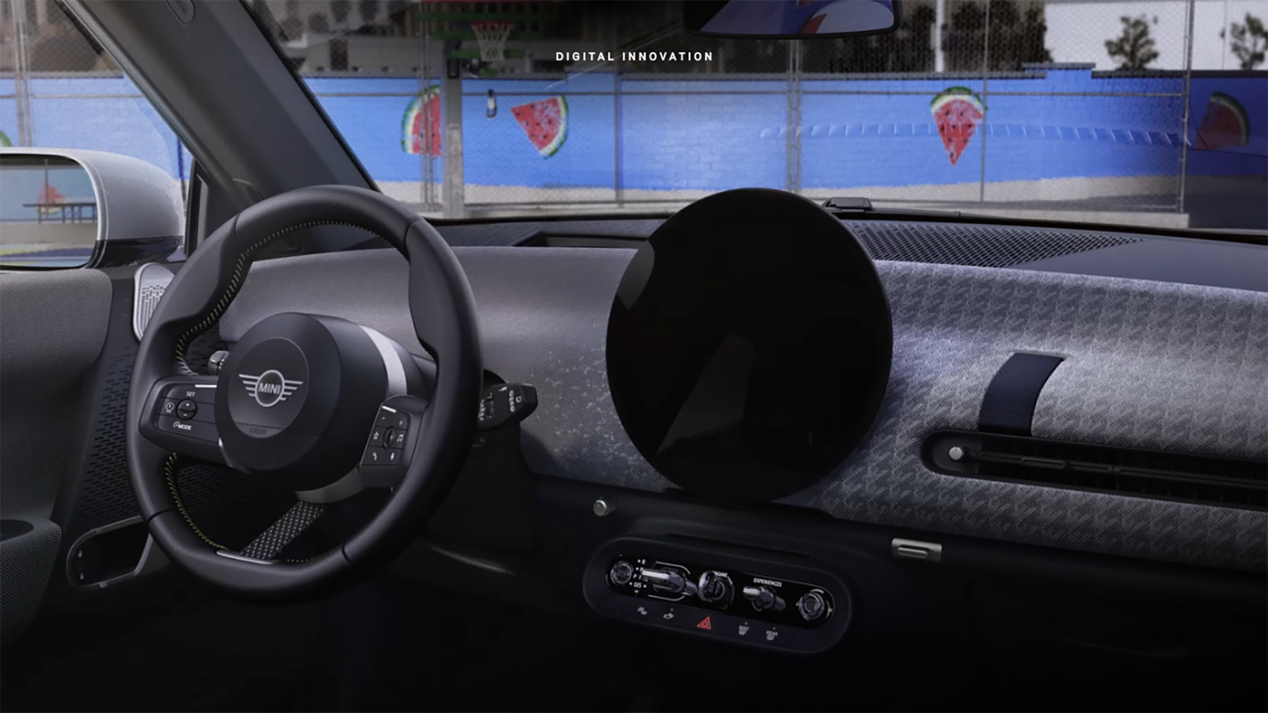 Closeup view of the MINI Cooper S 2 Door dashboard and 9.4 inch round OLED display from the perspective of the front
passenger's seat, with a purple wall including watermelons plastered on it visible through the windshield.