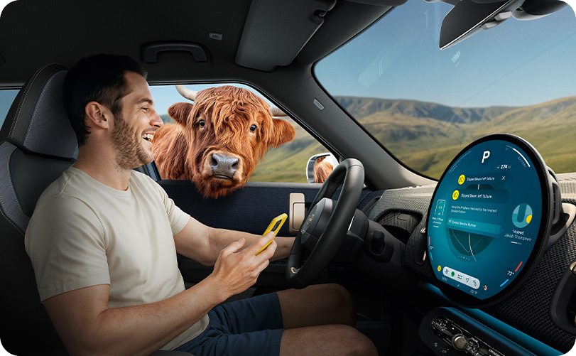 Highland cow looking through the window of a parked MINI Countryman S ALL4 at a driver holding a smartphone inside.
