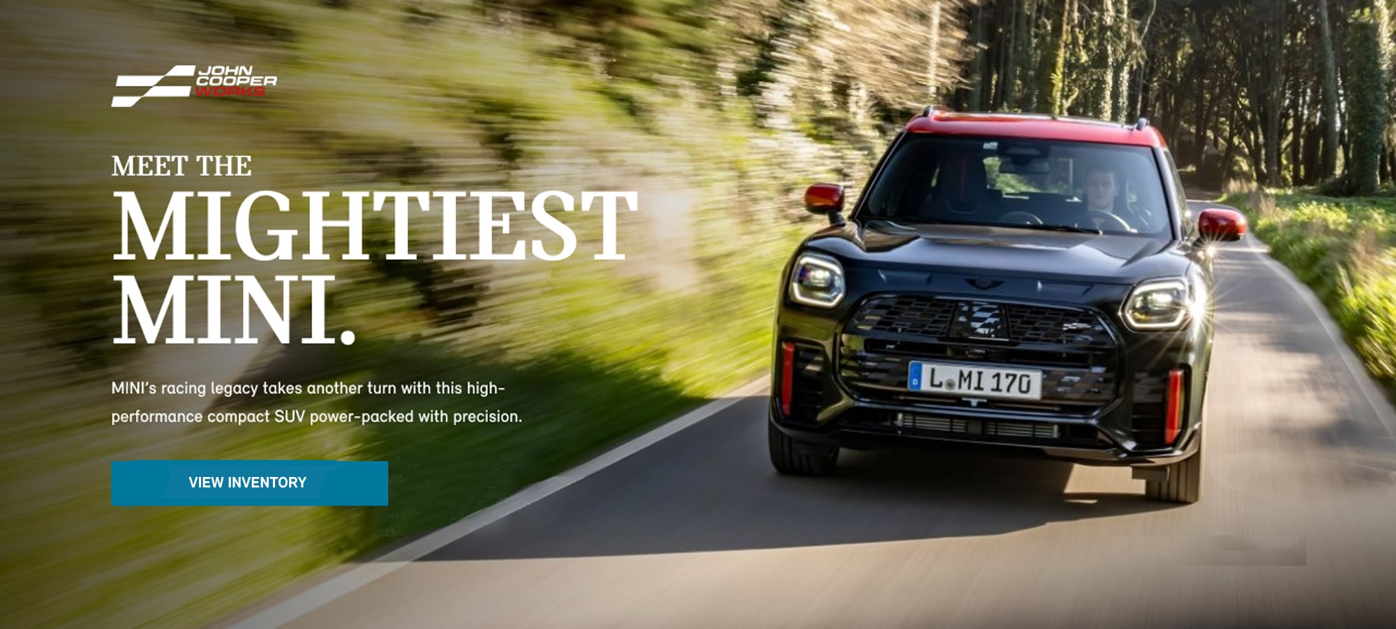 Front-view of a 2025 MINI JCW Countryman ALL4 driving down a road surrounded by blurred foliage.