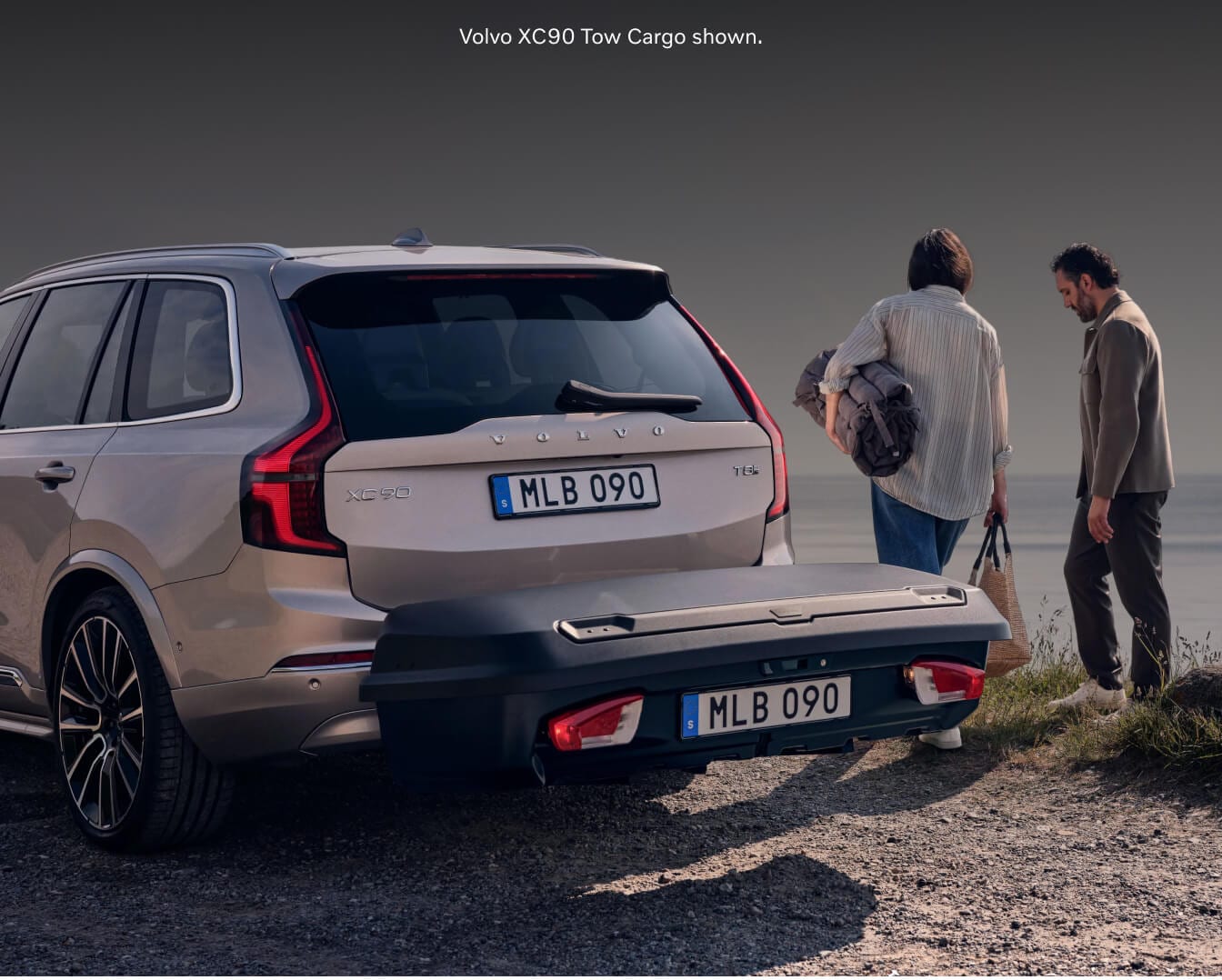 Rear view of a Sand Dune Volvo XC90 Ultra with a hitch-mounted tow cargo box by the sea.