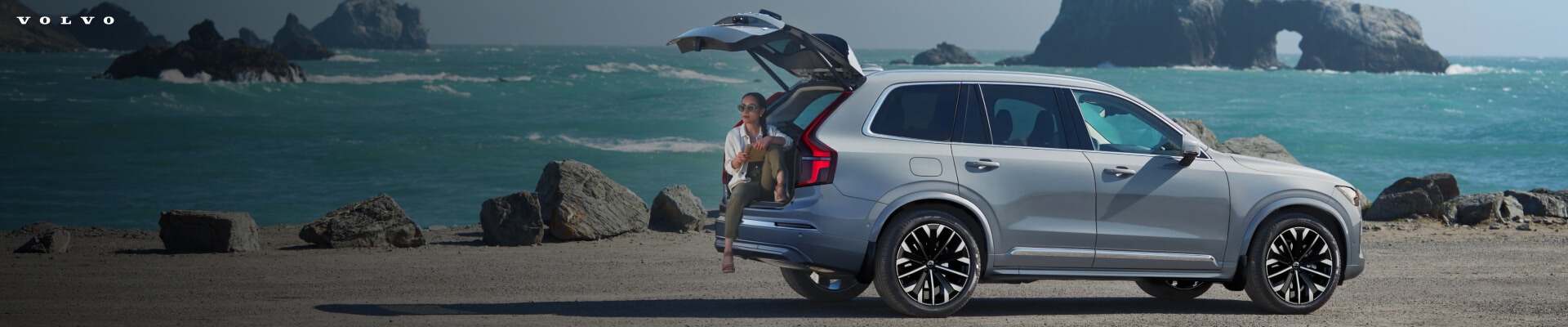 Rear-quarter view of a Vapour Grey Metallic Volvo XC90 Ultra with an open tailgate as a woman sits inside.