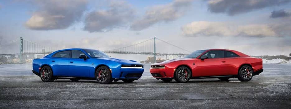 blue four-door and red two-door dodge chargers facing each other on a street in front of a bridge