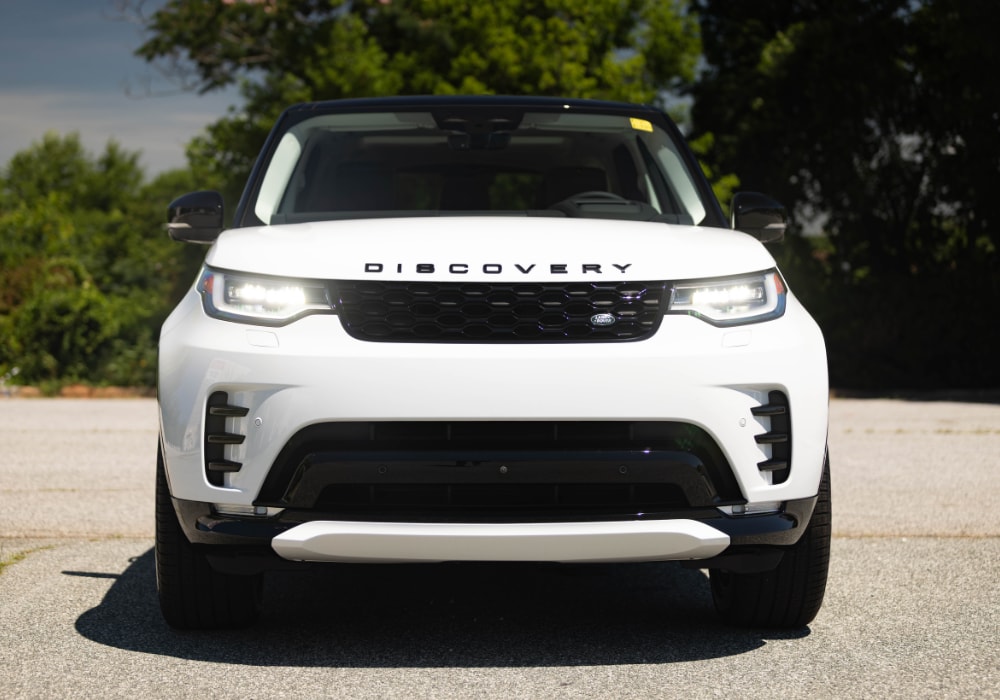 White 2025 Land Discovery SUV parked on a paved surface. The car's headlights are on and it is facing the camera