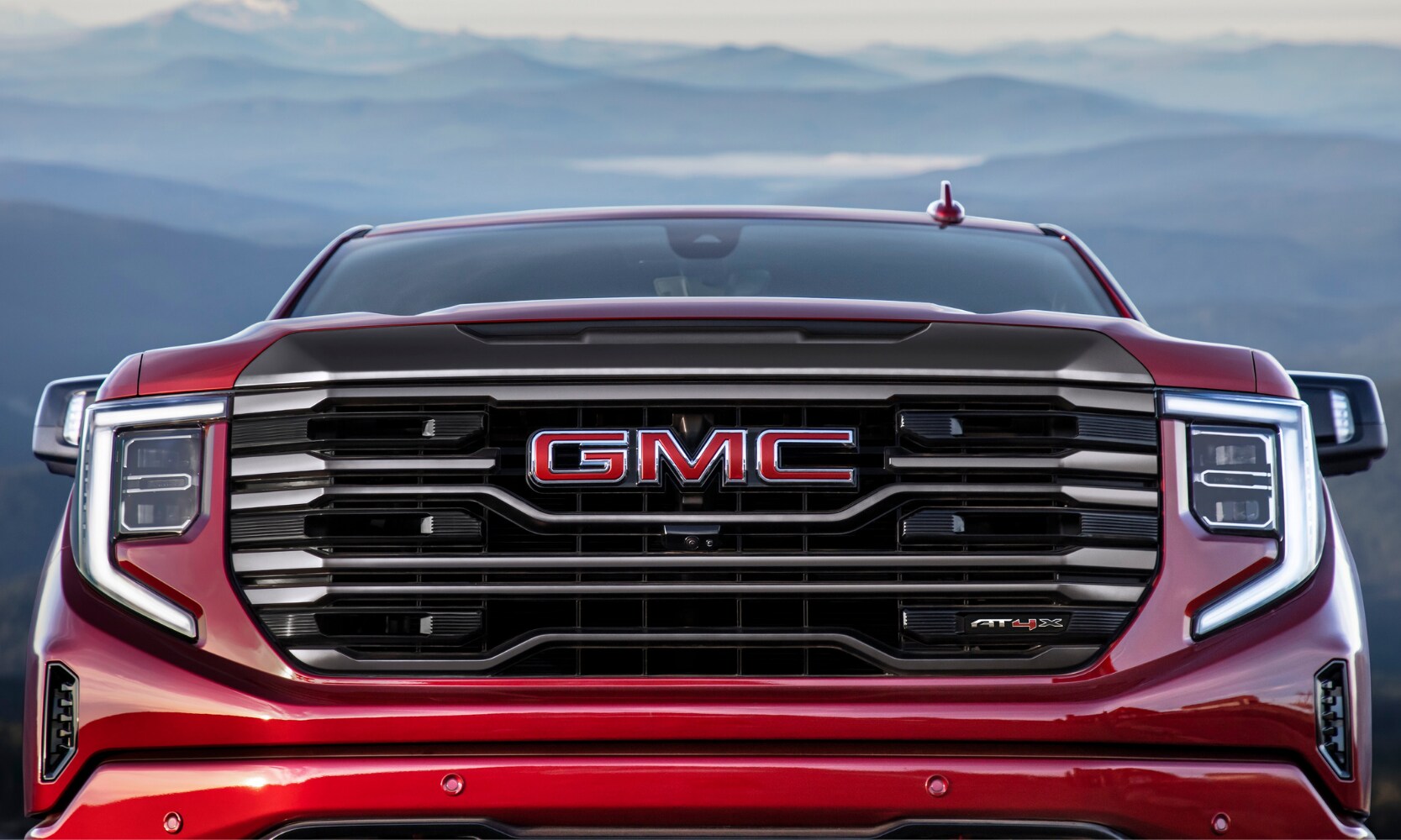 Close up view of the front grille on a red 2022 GMC Sierra 1500 AT4X off-road truck driving up to the camera lens overlooking a hazy distant moutain range