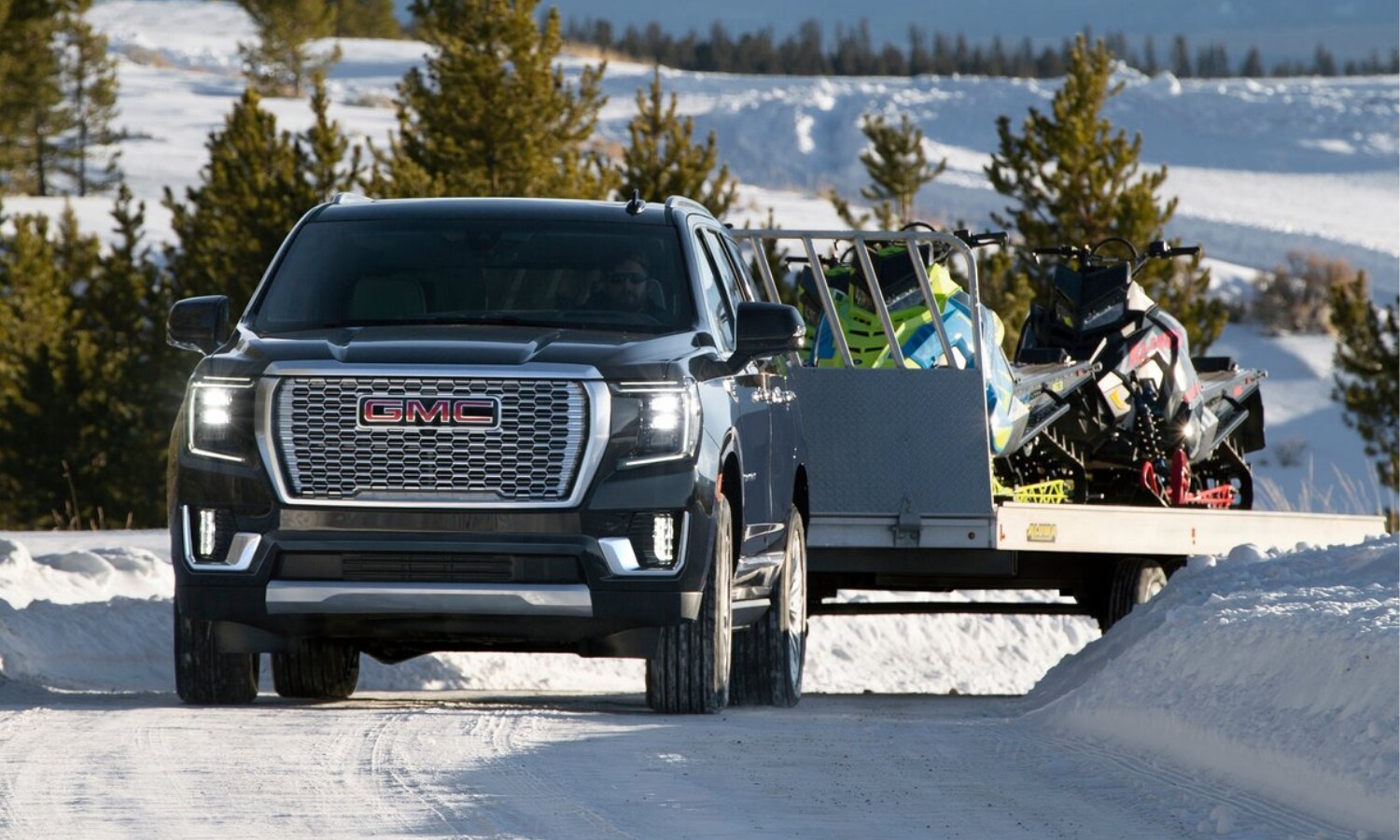 Black 2022 GMC Yukon Denali towing a large trailer loaded with snowmobiles through a snowy mountain pass