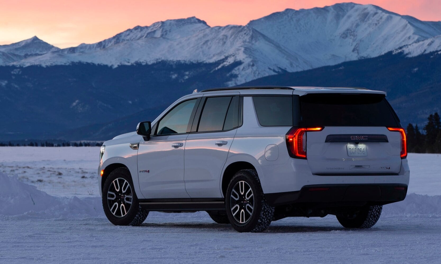 a white 2024 GMC Yukon parked in a victorious stance in a snowy clearing in front of snow-capped mountains at sunset with an orange-ish pink sky over the range