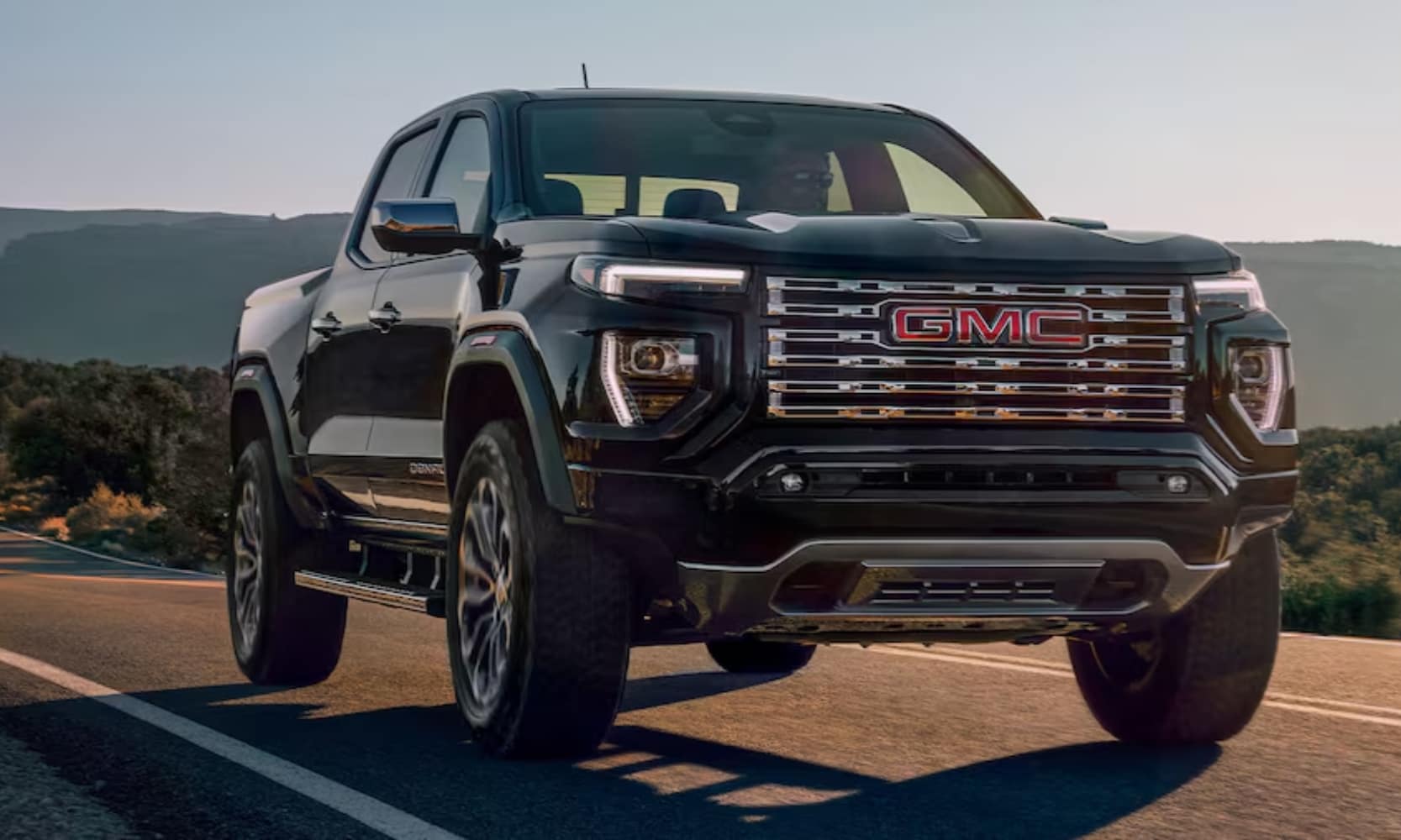 Exterior front image of a black 2024 GMC Canyon Denali driving up an open freeway with mountains in the background