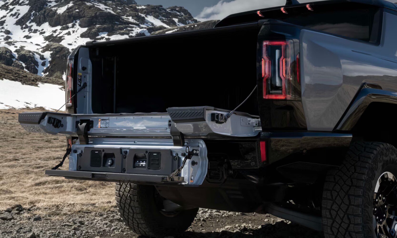 Closeup side angle image of a gray 2025 GMC Hummer EV Truck's MultiPro Tailgate opened in a snowy mountain landscape