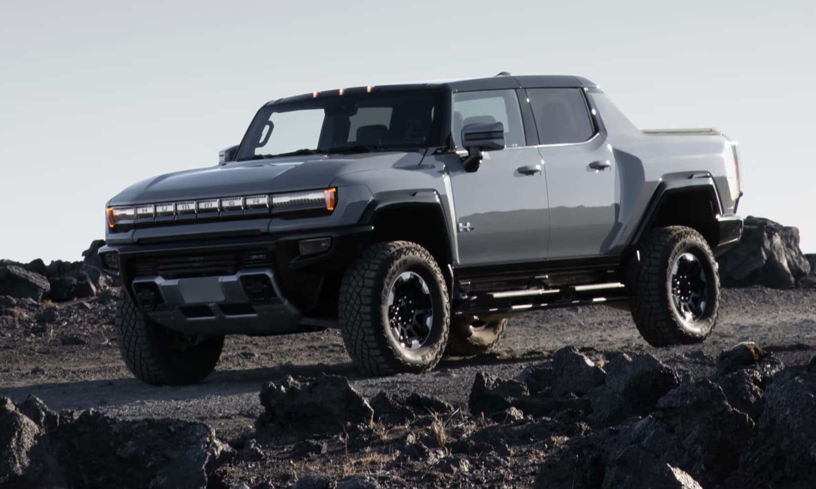 Exterior side angle image of a gray 2025 GMC Hummer EV Truck driving on a rocky mountain road