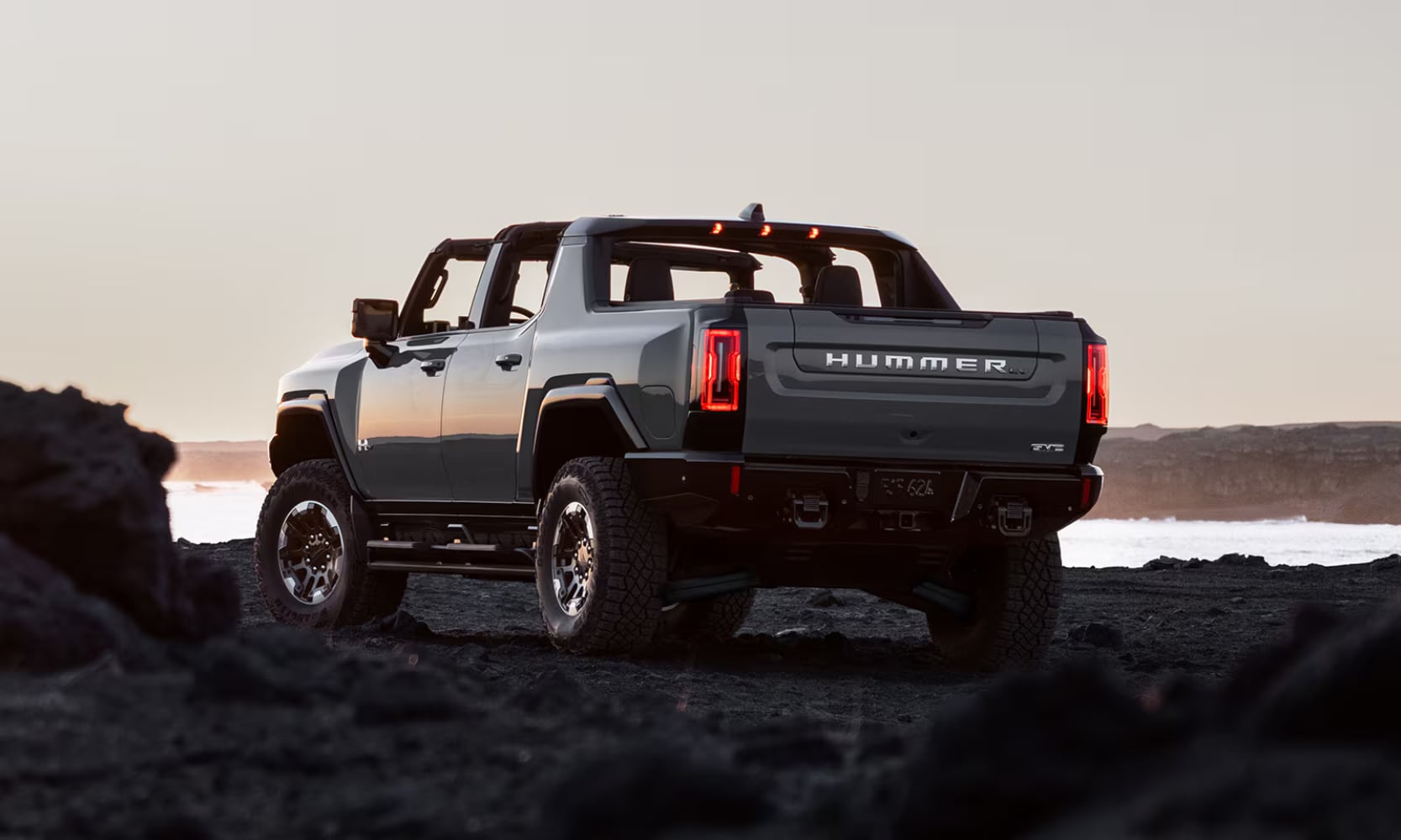 Exterior rear image of a gray 2026 GMC Hummer EV Pickup parked on a rocky water landscape with its roof panels removed