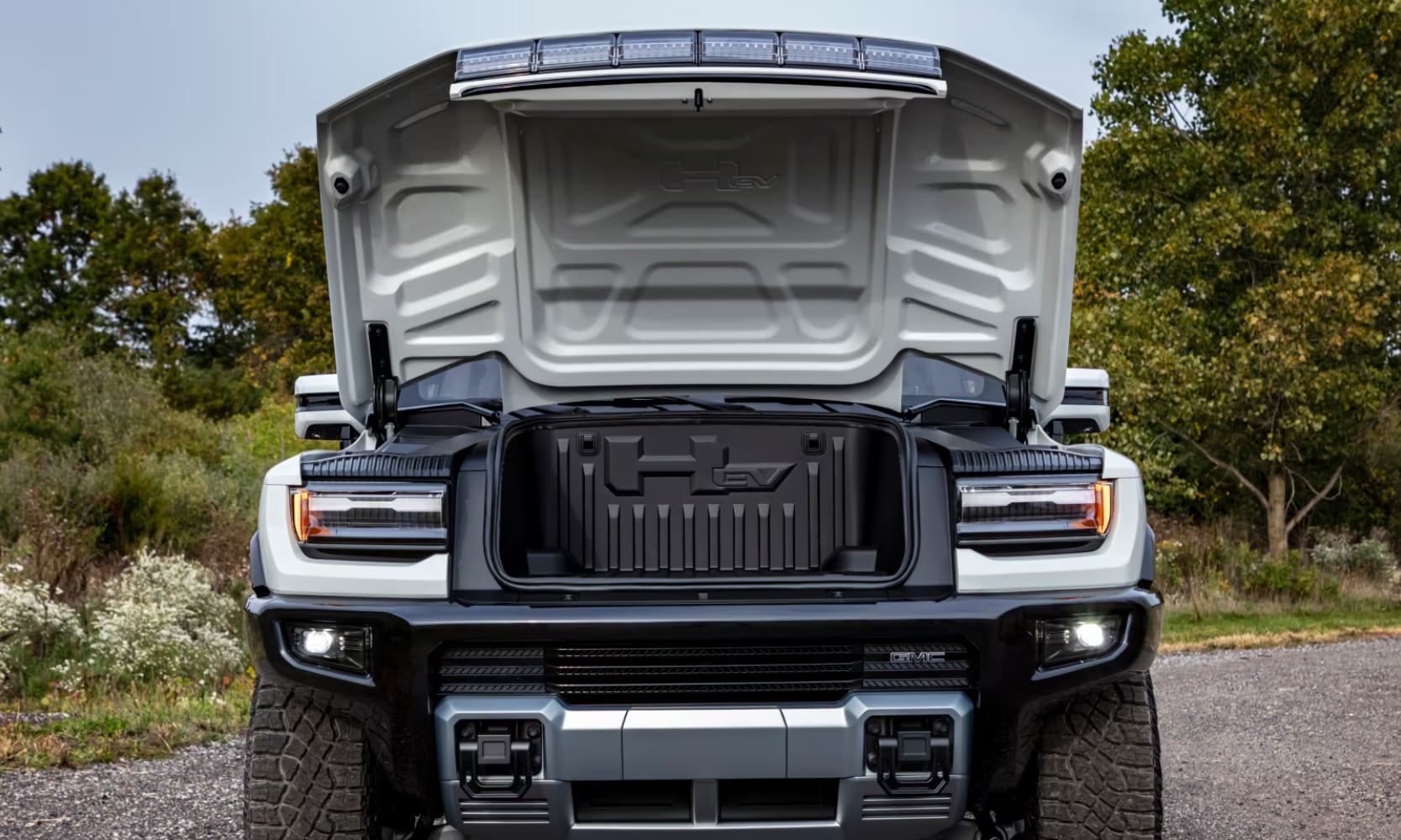 Front view exterior image of a white 2025 GMC Hummer EV Truck's front power eTrunk opened with trees behind it