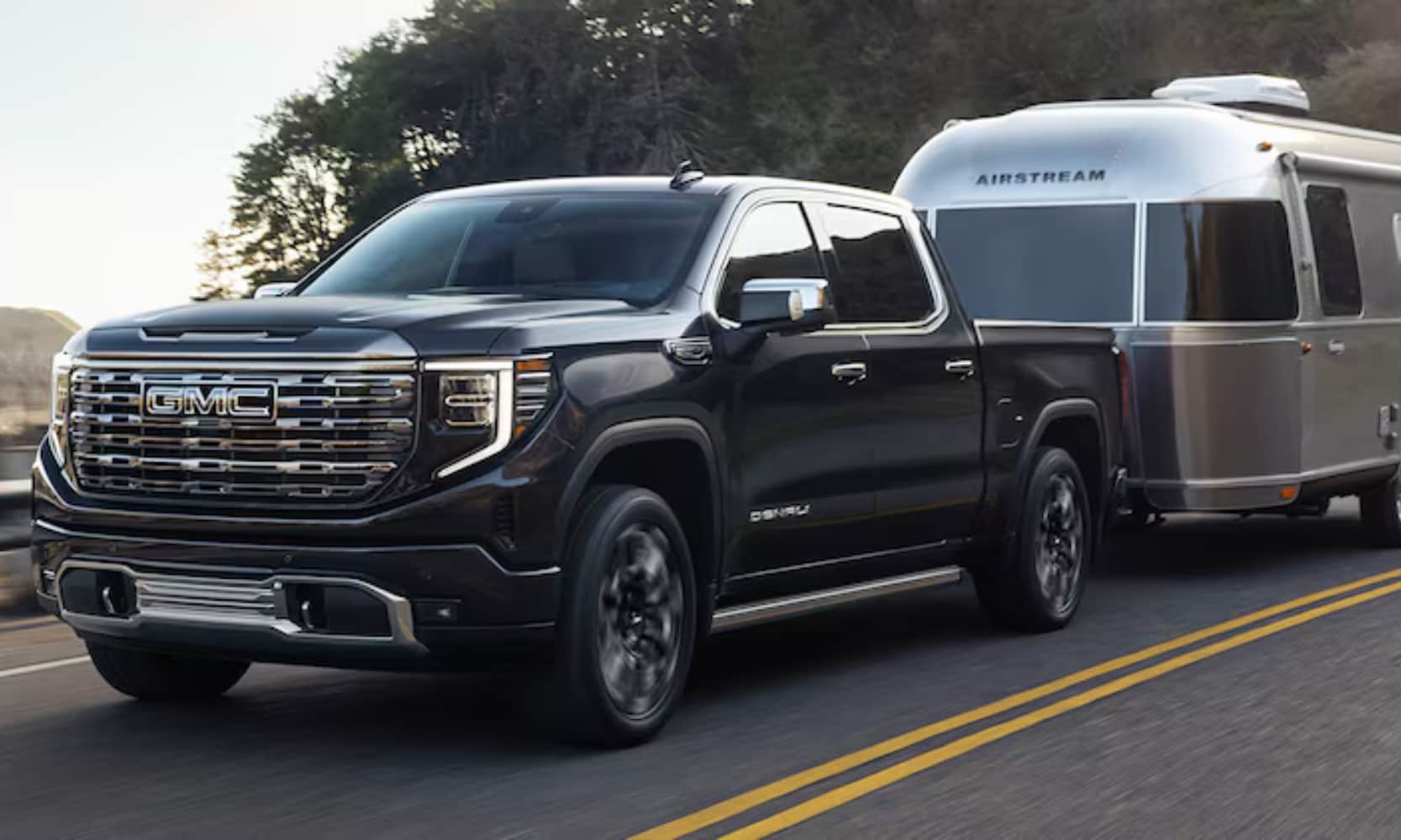 Exterior front side angle image of a black 2025 GMC Sierra 1500 Denali towing a silver camper on a freeway