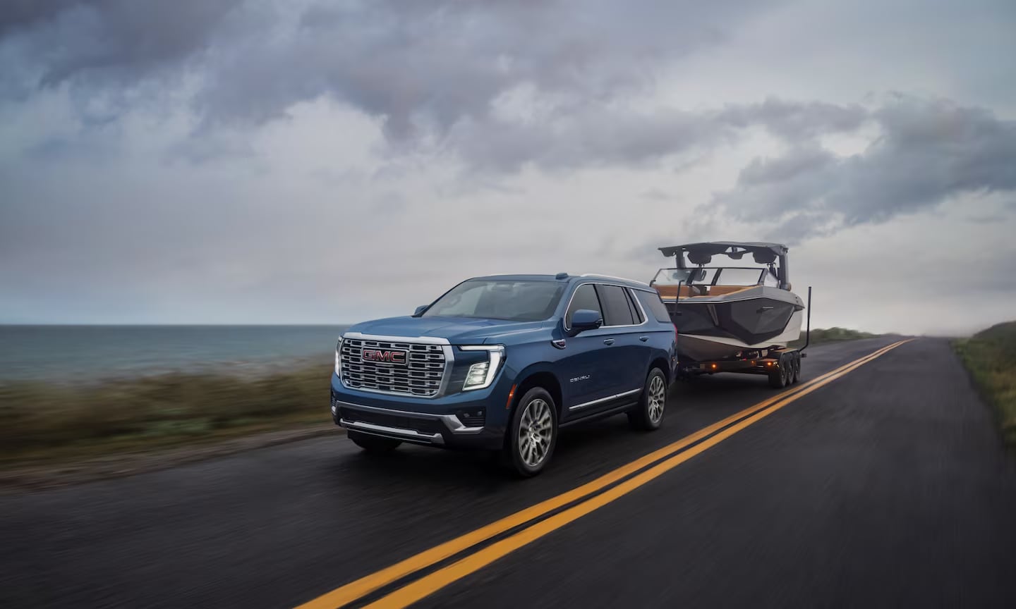 Exterior image of a blue 2026 GMC Yukon towing a small boat on a waterfront open freeway