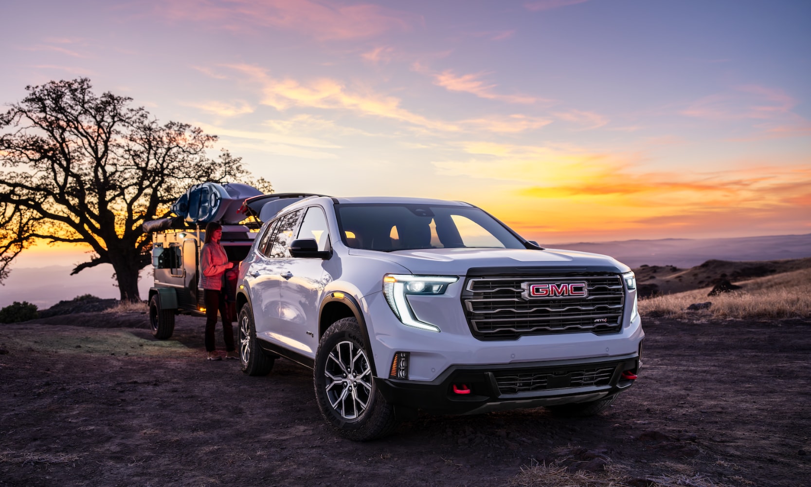 Exterior side image of a white 2024 GMC Acadia AT4 parked on a mountain with camping gear hauled