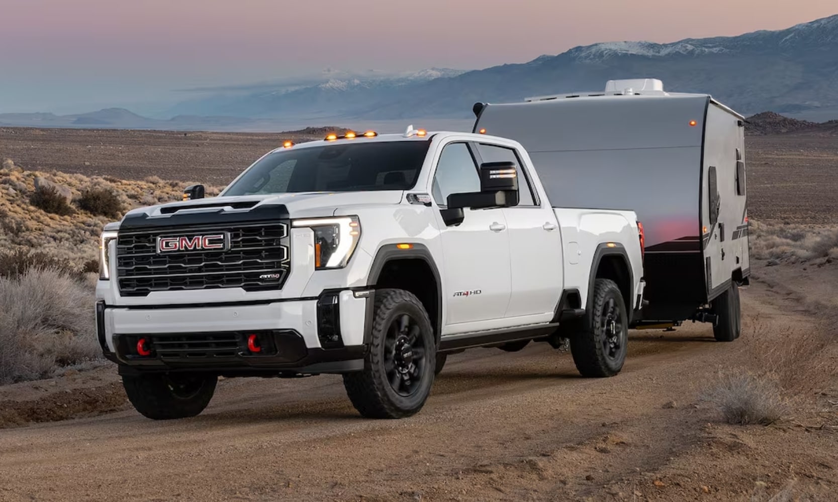 Exterior side angle image of a white 2025 GMC Sierra 2500 HD AT4 towing a small camper on a dirt road with mountains in the background