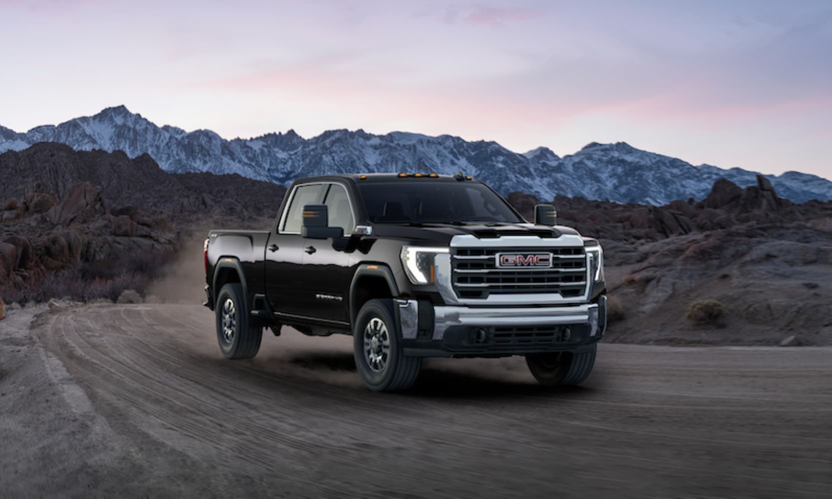 Exterior front angle mage of a black 2026 GMC Sierra 2500 HD SLE driving on an open freeway with mountains in the background