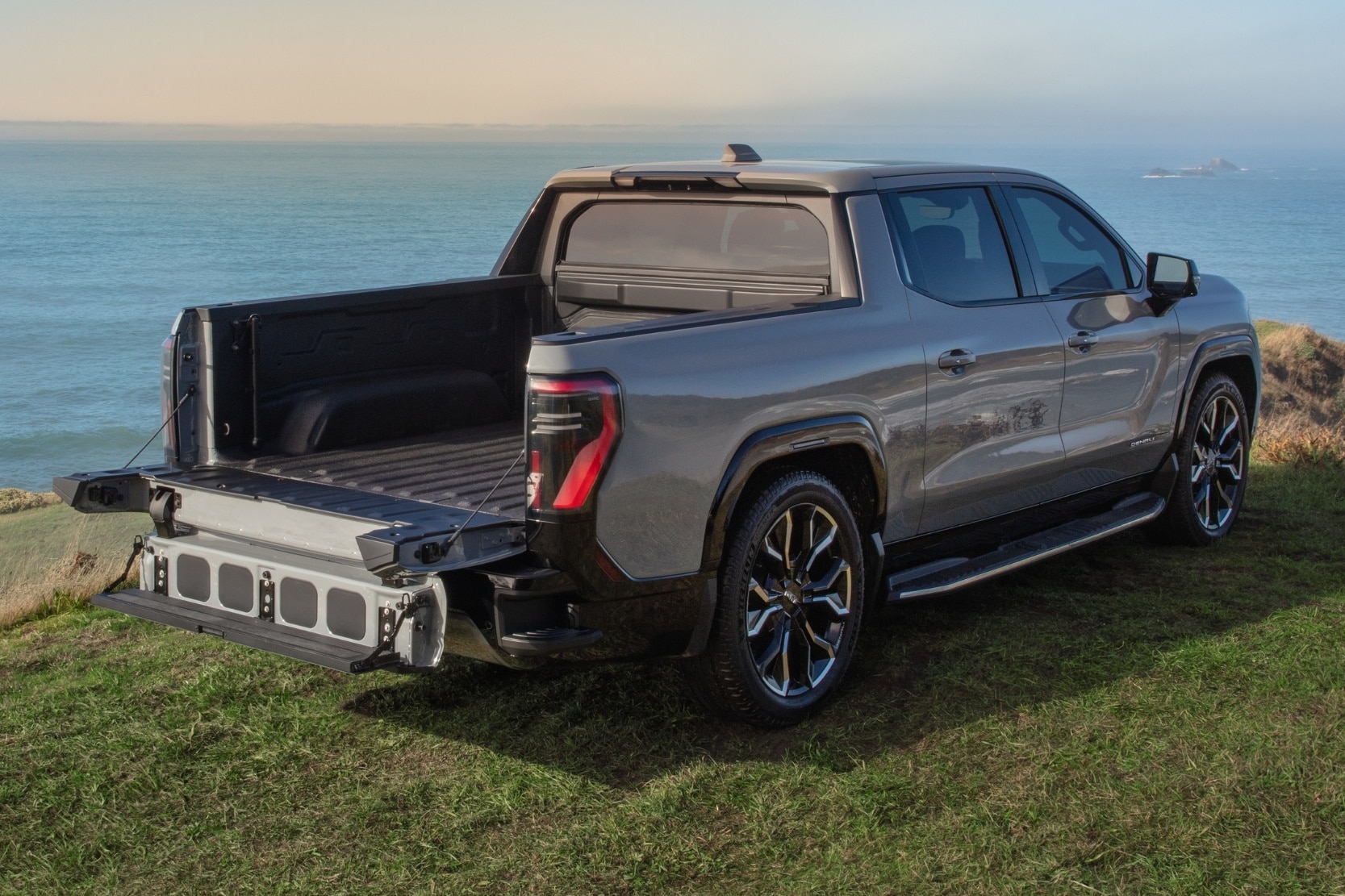 rear exterior view of the 2025 GMC Sierra EV Denali parked on the edge of a cliff overlooking the sea with the multipro functional tailgate opened