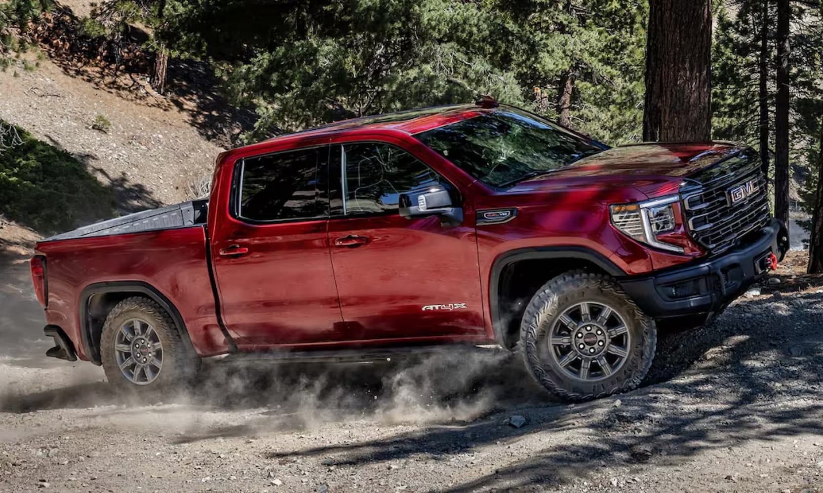 Exterior side image of a red 2025 GMC Sierra 1500 AT4X driving up a rocky hill in a forest landscape