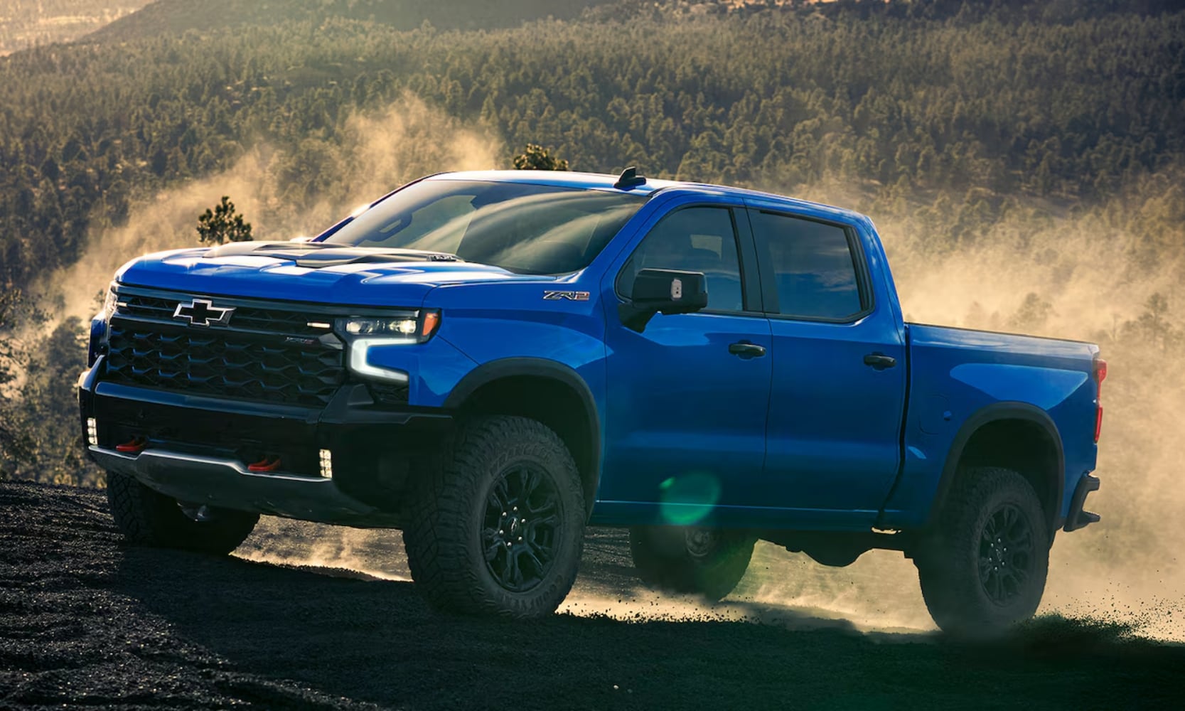 Exterior side angle image of a 2025 Chevy Silverado 1500 ZR2 driving up a dirt hill in a forest mountain landscape
