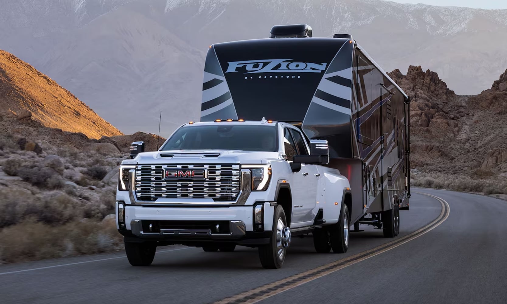 Exterior image of a white 2025 GMC Sierra 2500 HD towing a camping RV in a mountain landscape