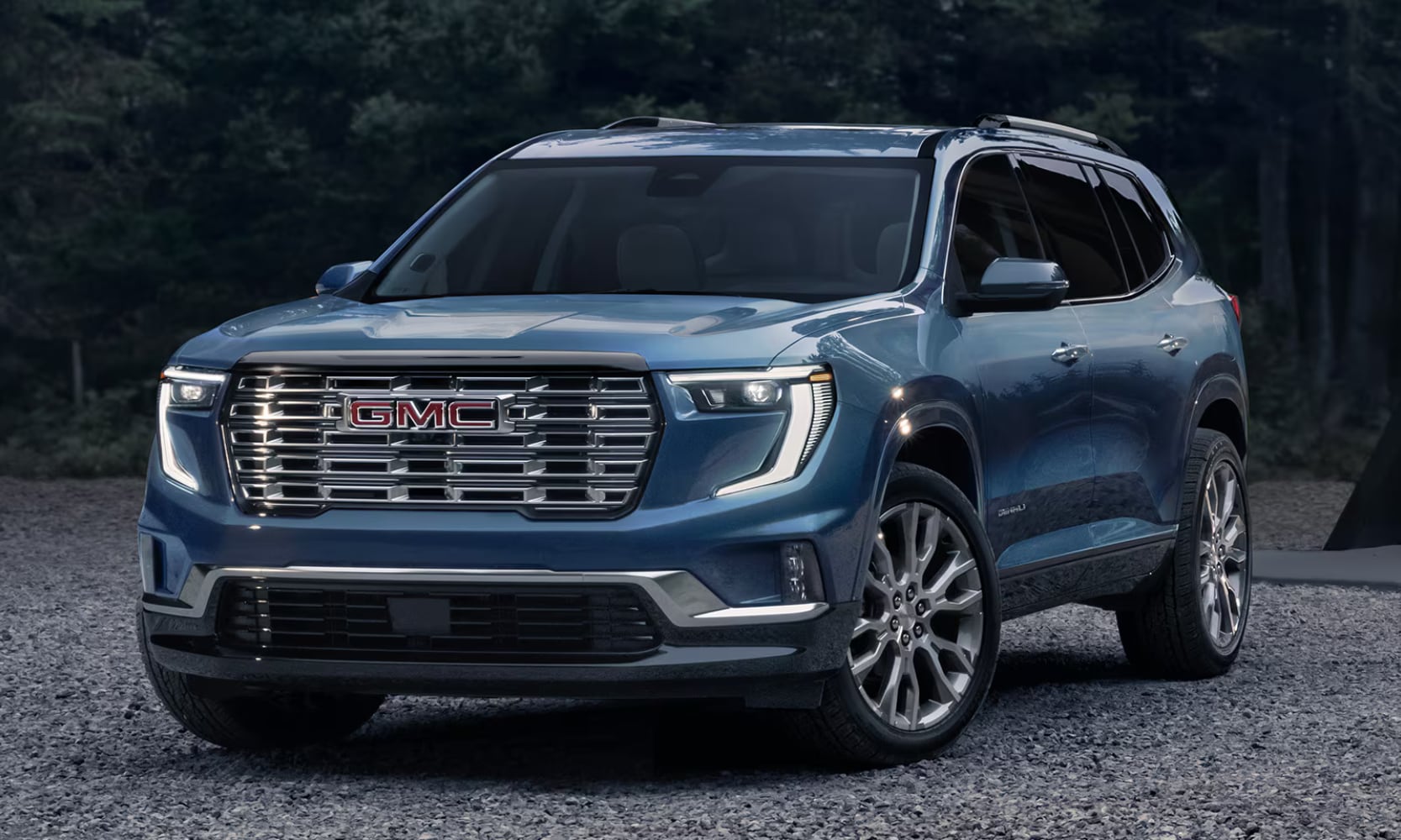 Exterior side angle image of a blue 2026 GMC Acadia Denali parked on a gravel driveway with forest background views