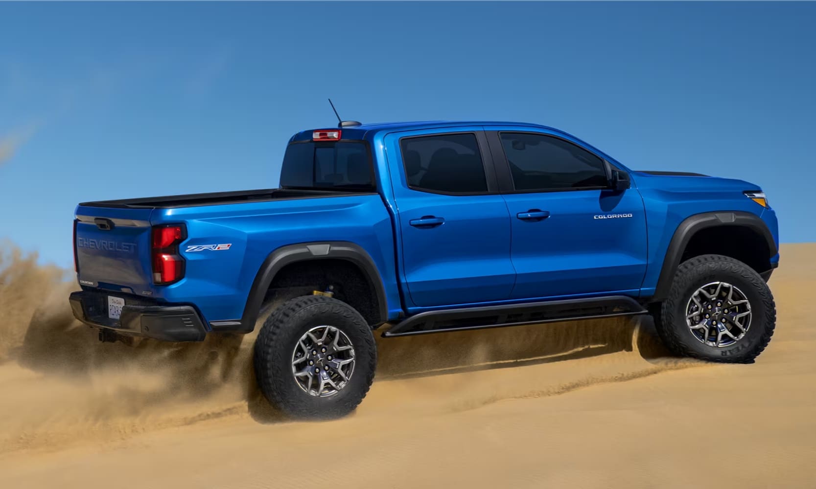 Exterior image of a blue 2024 Chevy Colorado ZR2 driving in a desert landscape with sand blowing
