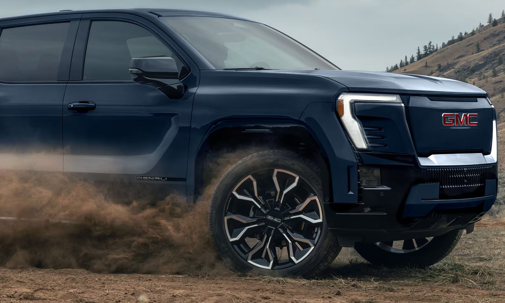 Exterior side image of a dark blue 2026 GMC Sierra EV Denali driving on a dirt road with dirt blowing behind the front wheels