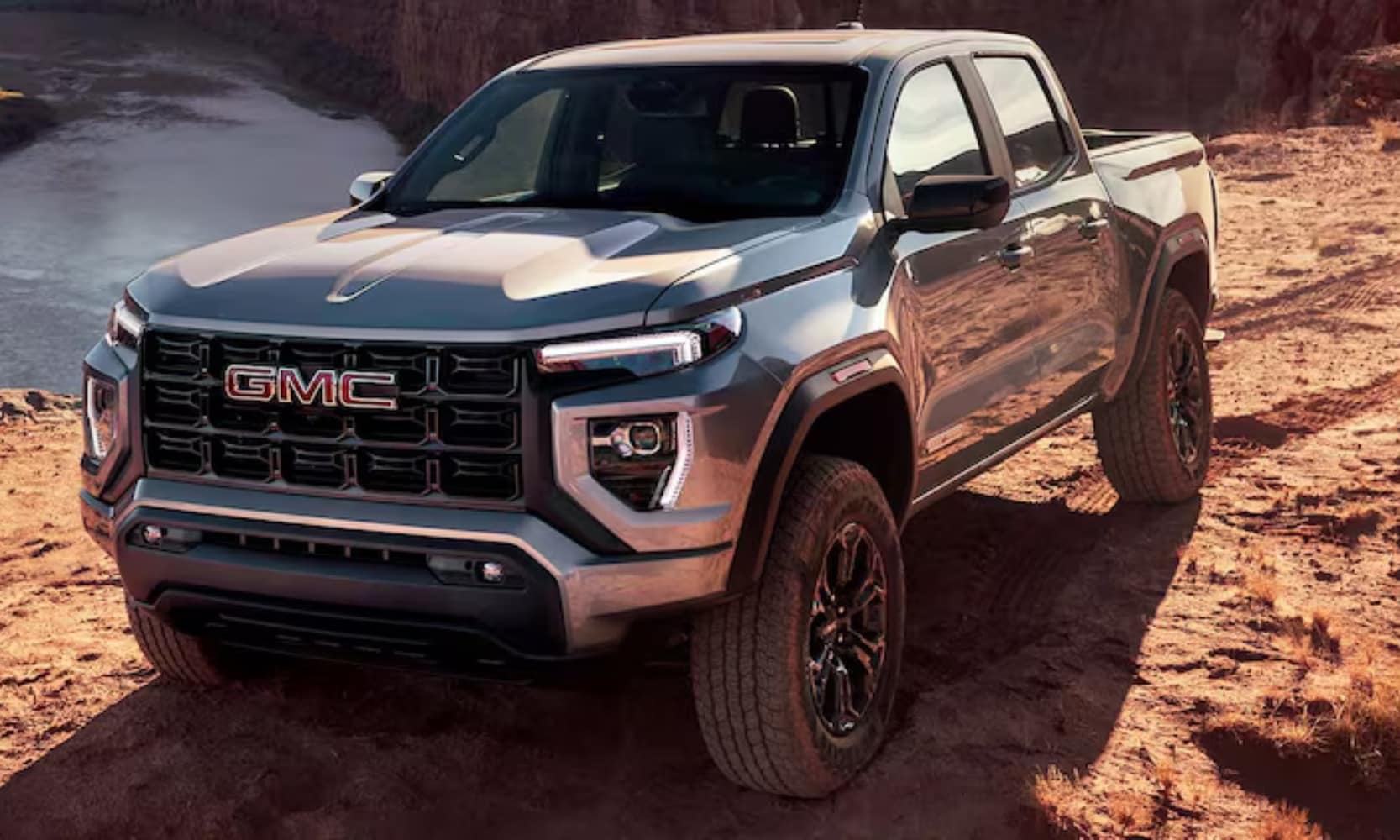 Exterior front angle image of a light gray 2024 GMC Canyon Elevation parked on a rocky hill with water in the background