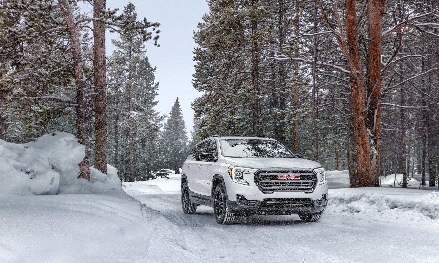 Exterior front image of a white 2024 GMC Terrain AT4 driving on a tree-lined snow-covered road on a snowy day