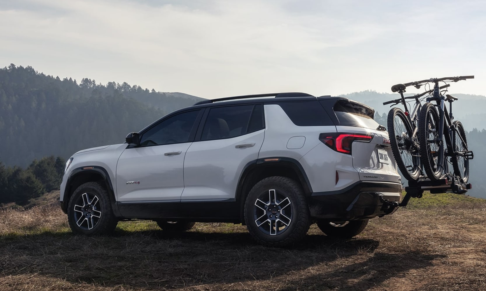 Exterior side image of a white 2026 GMC Terrain AT4 towing two mountain bikes in a mountain landscape