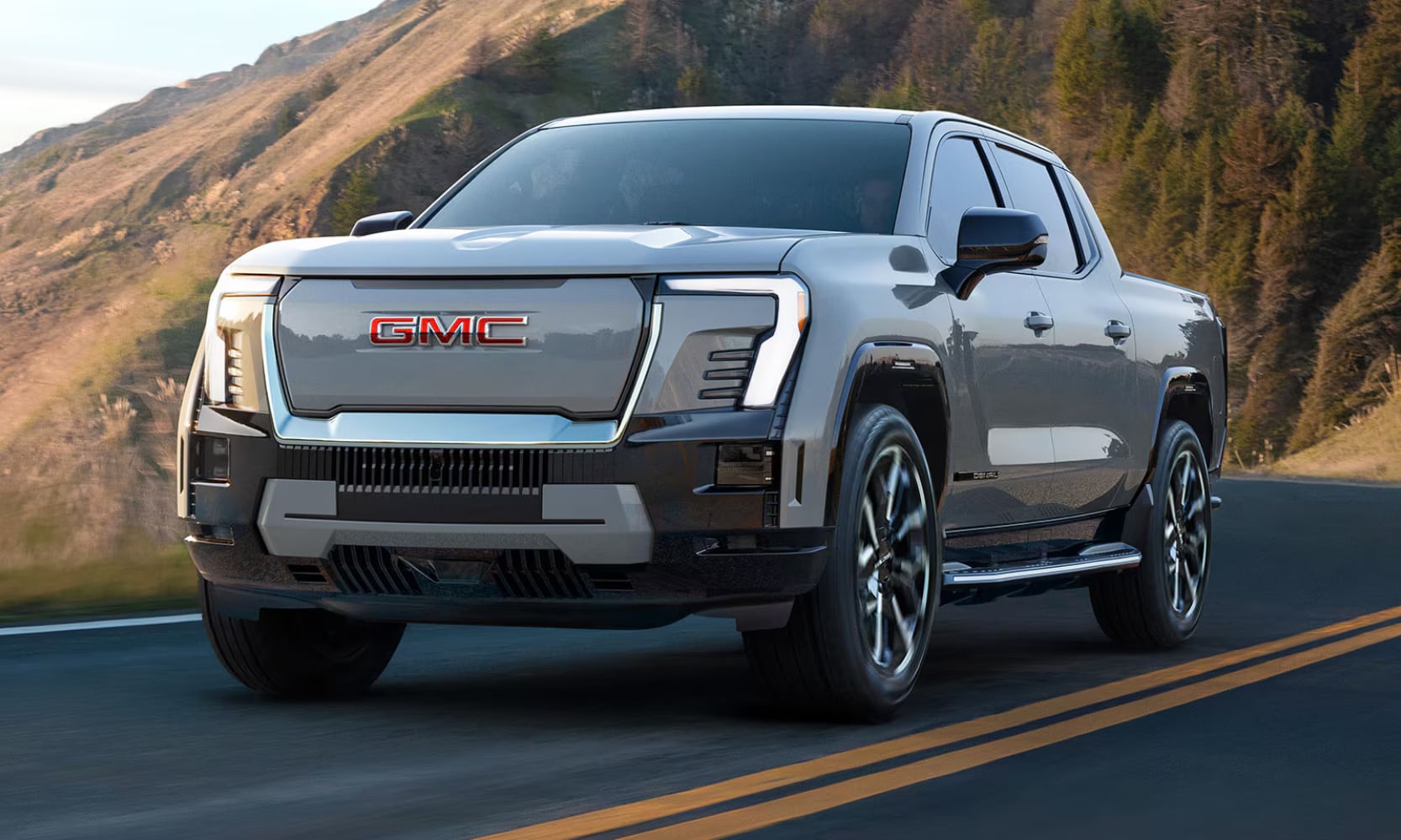 Exterior front angle image of a white 2026 GMC Sierra EV driving on a mountainous freeway