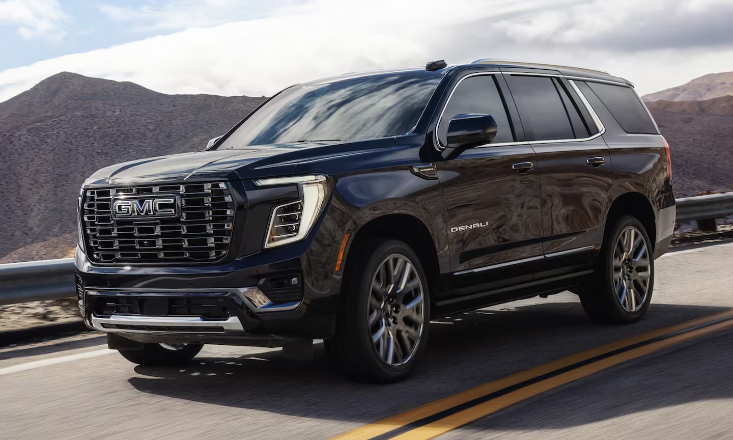 Exterior side image of a black 2026 GMC Yukon driving on an open mountainous freeway