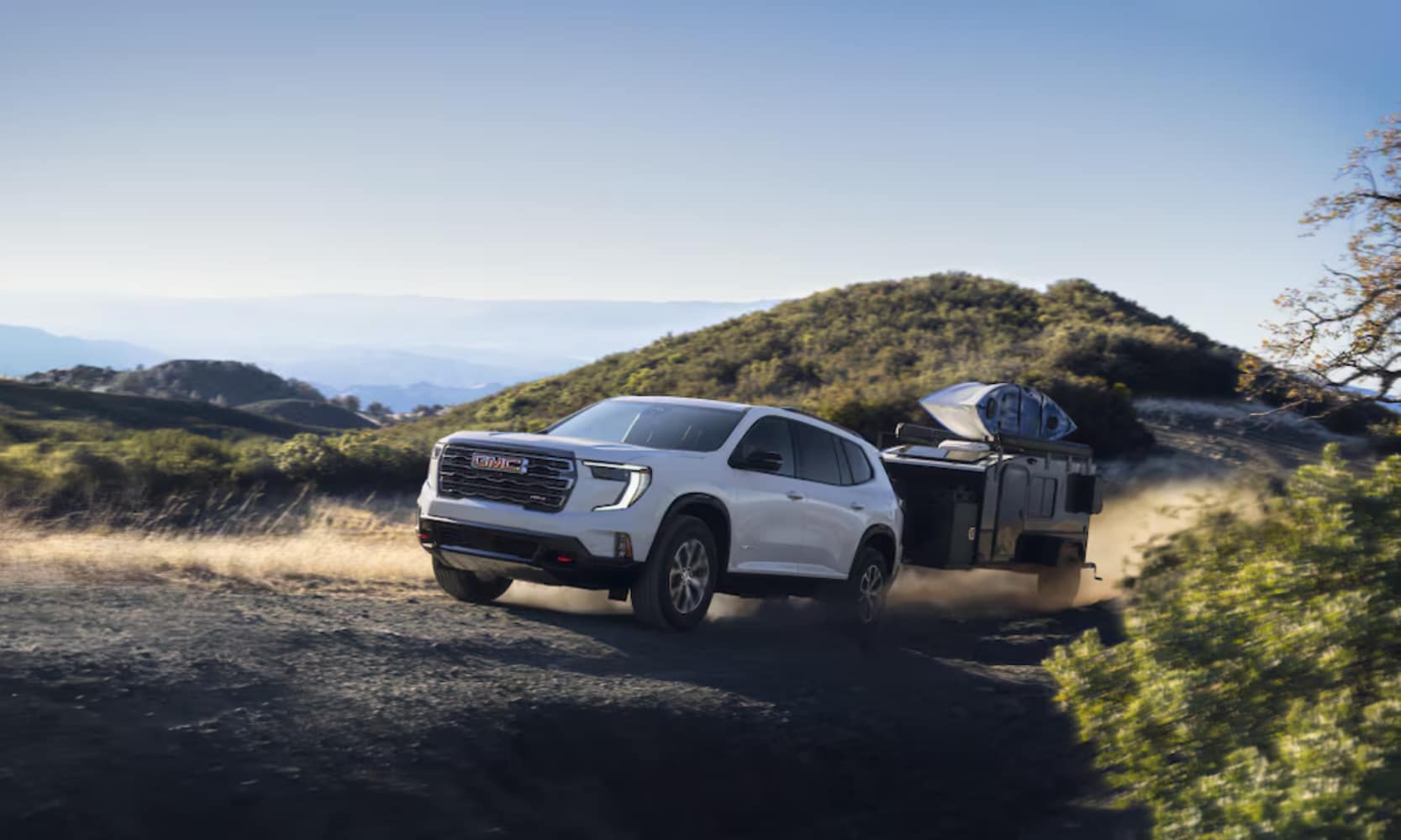Exterior image of a white 2025 GMC Acadia towing a small camper up a hill