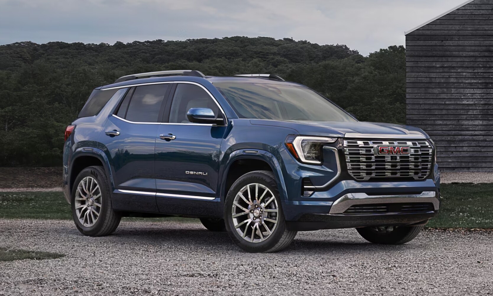 Exterior image of a blue 2026 GMC Terrain Denali parked outside on a gravel road with trees in the background