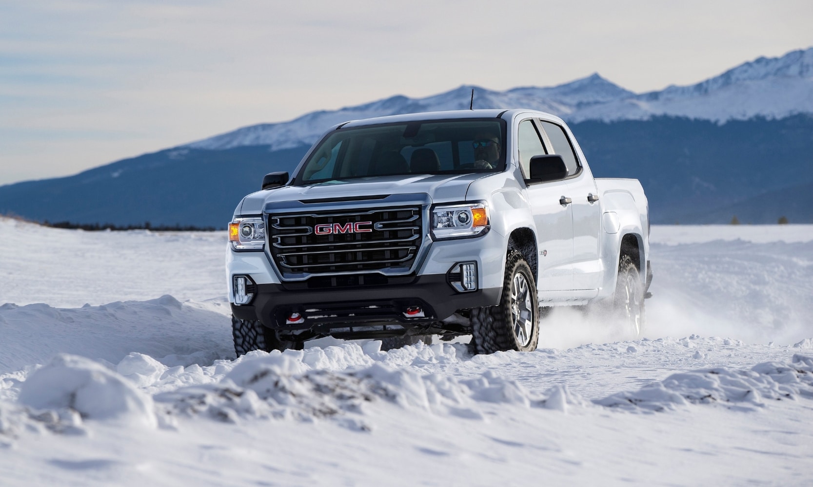 Exterior front image of a white 2021 GMC Canyon AT4 driving through deep snow with mountains in the background