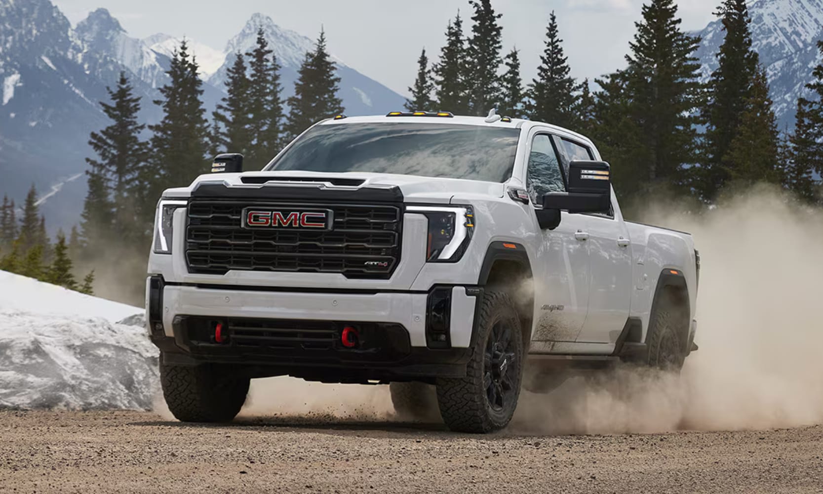 Exterior front image of a white 2026 GMC Sierra HD AT4 driving on a dirt road through a snowy mountain forest landscape
